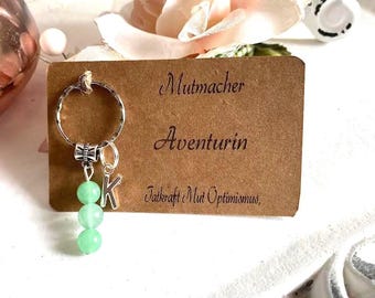 May include: A silver keychain with a green stone charm and the letter "K" hanging from it. The keychain is attached to a brown card with the words "Mutmacher Aventurin" printed on it.
