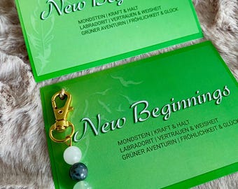 May include: A green card with the text "New Beginnings" and a list of gemstones and their associated meanings. The card is attached to a gold key chain with three beads: a white bead, a dark green bead, and a light green bead.