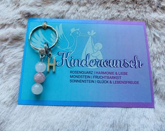 May include: A keychain with a silver ring, a gold letter "H" charm, and three pink and white gemstone beads. The keychain is displayed on a blue card with the German word "Kinderwunsch" and other text.