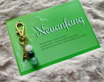 May include: A gold keychain with a charm that says "A" and has three green and white stones. The keychain is attached to a green card with the text "Neuanfang" and a list of gemstones and their meanings.