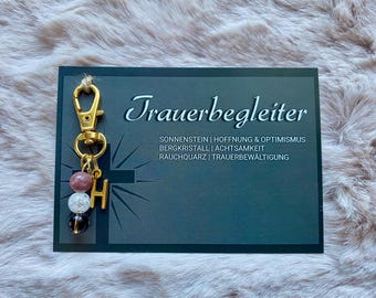May include: Gold-toned keychain with a charm featuring a letter H and a variety of small stones. The keychain is labeled "Trauerbegleiter" with the text "Sonnenstein | Hoffnung & Optimismus Bergkristall | Achtsamkeit Rauchquarz | Trauerbewältigung".