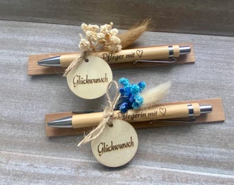 May include: Two wooden pens with silver accents, each resting on a small wooden base. The pens are adorned with dried flowers and a small wooden tag that says "Glückwunsch". One pen says "Pfleger mit ♡" and the other says "Pflegerin mit ♡".