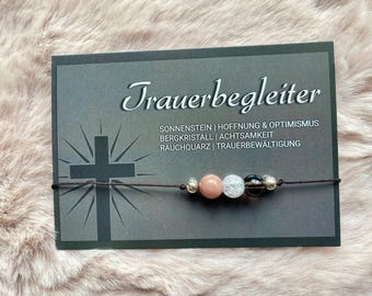 May include: A black card with the text "Trauerbegleiter" and the words "Sonnenstein | Hoffnung & Optimismus Bergkristall | Achtsamkeit Rauchquarz | Trauerbewältigung" printed on it. A brown string with beads is attached to the card.