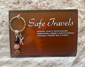 May include: A silver key chain with a black obsidian, pink rose quartz, and tiger's eye stone charm. The charm is attached to a silver letter K. The key chain is displayed on a card that reads "Safe Travels" and lists the properties of each stone.