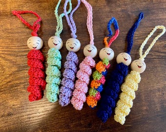 May include: A collection of colorful, handmade crochet ornaments. Each ornament features a wooden bead with a smiling face and a looped string for hanging. The crochet sections are in various colors, including red, green, purple, pink, orange, blue, and yellow.