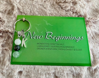 May include: A green card with the text "New Beginnings" and a list of gemstones and their meanings. A silver key chain with a letter K charm and three beads: white, black, and green.
