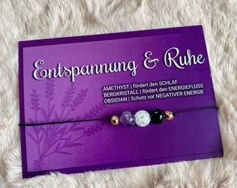 May include: A purple card with the text "Entspannung & Ruhe" and a black string with amethyst, clear quartz, and black obsidian beads. The text below the title reads "Amethyst | fördert den Schlaf, Bergkristall | fördert den Energiefluss, Obsidian | Schutz vor negativer Energie."