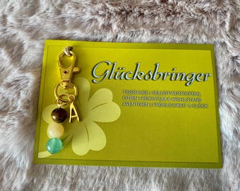 May include: Gold keychain with a charm featuring the letter "A" and various gemstones, including tiger's eye, citrine, and aventurine. The card reads "Glücksbringer" and lists the meanings of the stones: Tiger's eye for self-esteem, citrine for wealth and prosperity, and aventurine for happiness and luck.