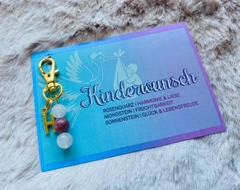 May include: A gold key chain with a charm that says "H" and three beads. The beads are white, pink, and white. The key chain is attached to a card that says "Kinderwunsch" with the text "Rosenquarz | Harmonie & Liebe Mondstein | Fruchtbarkeit Sonnenstein | Glück & Lebensfreude".