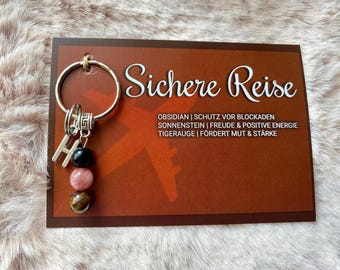 May include: A keychain with a silver ring, an initial "H" charm, and three gemstone beads in black, pink, and brown. The keychain hangs on a brown card with the words "Sichere Reise" and other text.