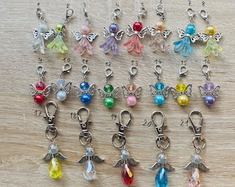 May include: A collection of angel-themed keychains with silver-tone wings and lobster clasps. Each keychain features a colorful bead and a crystal-like body in various colors, including yellow, blue, pink, and purple. The keychains are arranged on a light wood surface.