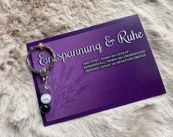 May include: A purple card with the text "Entspannung & Ruhe" and a list of crystals and their properties. A silver key chain with three beads: amethyst, clear quartz, and black obsidian.