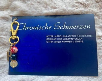 May include: A gold keychain with three beads: red with white floral pattern, black, and clear with gold flecks. The keychain is on a blue card with the text "Chronische Schmerzen" and information about the stones: "ROTER JASPIS | löst ÄNGSTE & SCHMERZEN OBSIDIAN | löst VERSPANNUNGEN CITRIN | gegen KUMMER & STRESS"