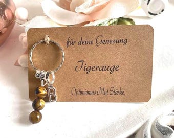 May include: A keychain with a silver ring, a letter "B" charm, and three tiger eye beads. The keychain is attached to a brown card with German text that reads "für deine Genesung Tigerauge Optimismus Mut Stärke."