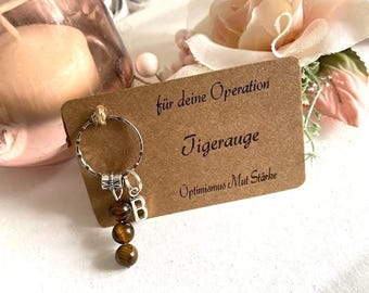 May include: A keychain with a silver ring, a bow charm, and three tiger eye beads. A small brown card with the text "für deine Operation Tigerauge Optimismus Mut Stärke" is in the background. A candle and flowers are also visible.