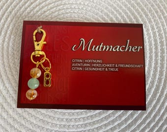 May include: A gold keychain with a charm that says "B" and has three small, colorful beads. The keychain is attached to a card that says "Mutmacher" and lists the stones and their meanings: Citrin | Hoffnung, Aventurin | Herzlichkeit & Freundschaft, Citrin | Gesundheit & Treue.