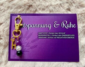 May include: A gold key chain with purple, clear, and black beads. The card behind the keychain reads "Entspannung & Ruhe" and lists the properties of amethyst, rock crystal, and obsidian.