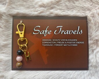 May include: A gold-colored keychain with a lobster clasp, three beads, and a letter "H" charm. The beads are black, pink, and brown. The keychain hangs from a brown card with the words "Safe Travels" and text in German.