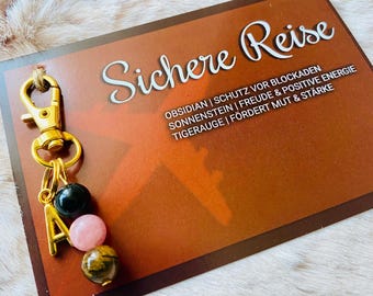 May include: A gold-colored keychain with the letter "A" and three gemstone beads: black, pink, and tiger's eye. The keychain hangs from a brown card with the words "Sichere Reise" and other text in German.