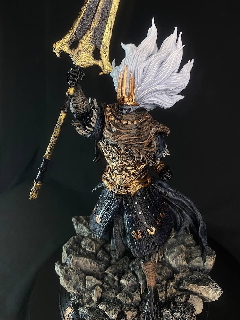 May include: A detailed figurine of a character from the video game Dark Souls. The figure is wearing a golden armor with a black cape and a white crown. The figure is holding a large golden spear with a black handle. The figure is standing on a rocky base.