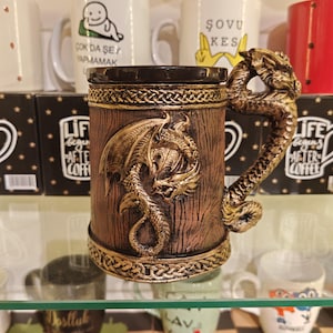 May include: A brown and gold dragon-themed mug with a detailed dragon handle and a relief carving of a dragon on the front. The mug has a wooden texture and is displayed on a glass shelf with other mugs in the background.