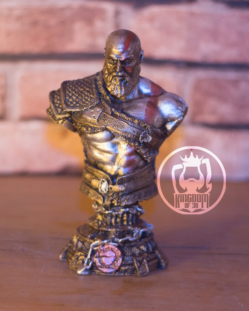May include: A detailed bust of a muscular, bearded figure with a red painted stripe on his head and face. The figure wears ornate armor and has a stern expression. The bust is on a decorative base with the text "KINGDOM OF 3D".