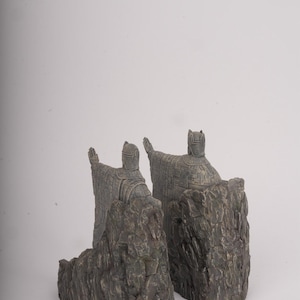 May include: A pair of bookends featuring figures in traditional robes with pointed headgear, each standing atop a textured, dark gray rock formation. The figures are depicted with arms extended outward, showcasing detailed fabric patterns and weathered stone textures.