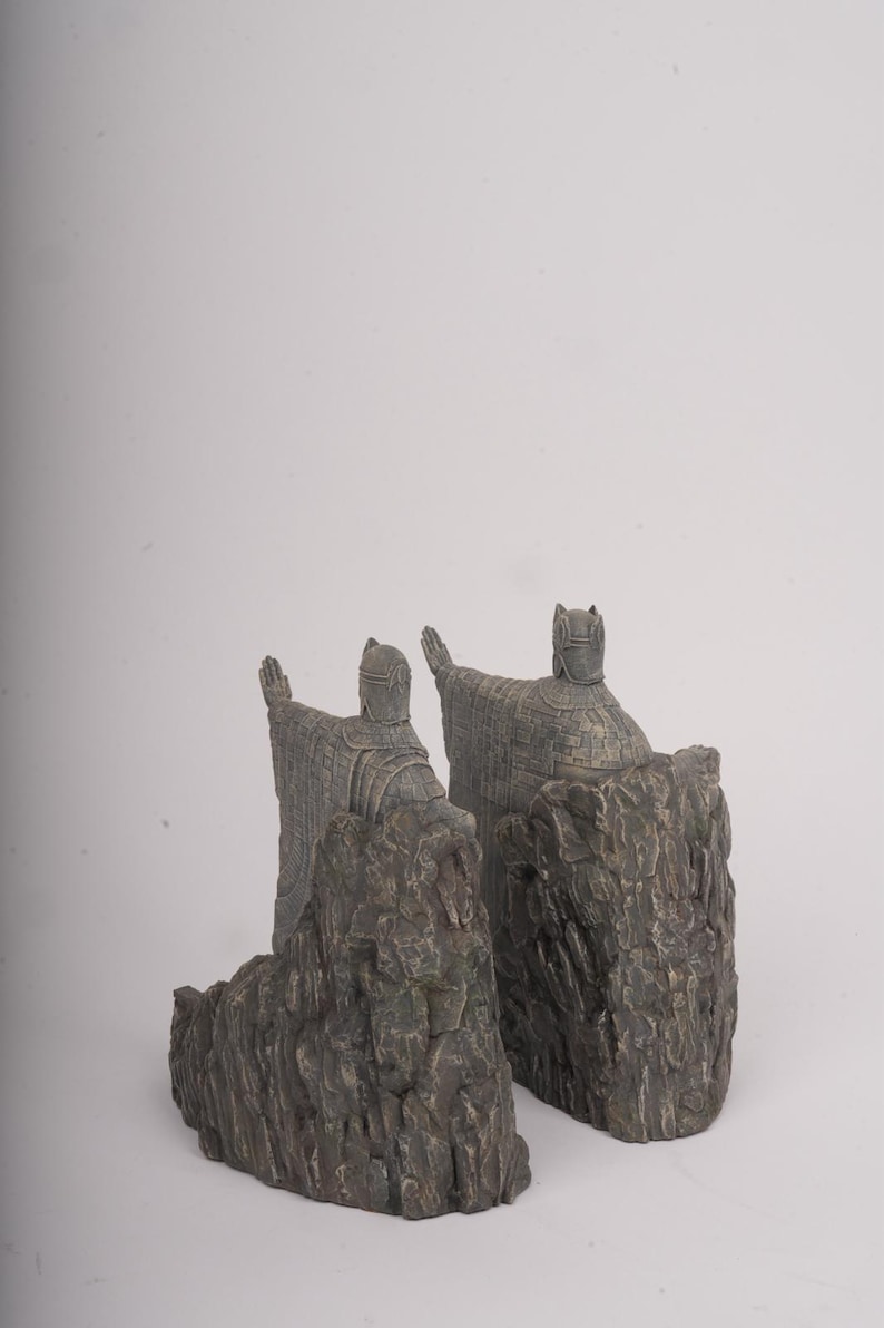 May include: A pair of bookends featuring figures in traditional robes with pointed headgear, each standing atop a textured, dark gray rock formation. The figures are depicted with arms extended outward, showcasing detailed fabric patterns and weathered stone textures.
