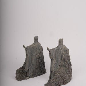 May include: A pair of grey bookends featuring standing figures in long robes and helmets, each with one arm extended forward. The figures are integrated into a textured, rocky base design, crafted with a weathered finish and detailed vertical lines.