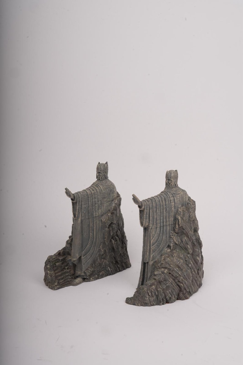 May include: A pair of grey bookends featuring standing figures in long robes and helmets, each with one arm extended forward. The figures are integrated into a textured, rocky base design, crafted with a weathered finish and detailed vertical lines.
