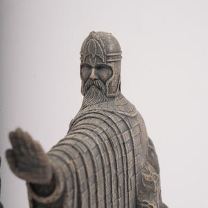 May include: A grey, stone-textured figurine of a bearded warrior wearing a detailed helmet and long, pleated robes. The figure is depicted with one arm extended forward, palm open, and stands against a plain, light-colored background.
