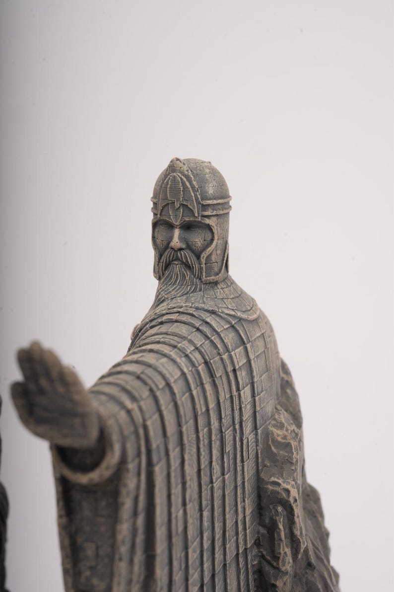 May include: A grey, stone-textured figurine of a bearded warrior wearing a detailed helmet and long, pleated robes. The figure is depicted with one arm extended forward, palm open, and stands against a plain, light-colored background.