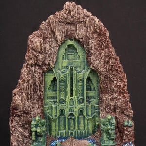 May include: A detailed miniature fantasy diorama featuring a green arched structure within a brown rock formation. The structure has intricate architectural details and is flanked by green statues. A blue stream flows at the base, creating a captivating scene.