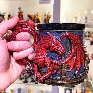 Könnte beinhalten: Eine rote Drachentasse mit passendem Griff. Die Tasse zeigt ein geformtes Drachendesign mit blauem Hintergrund und goldenen Akzenten. Die Tasse wird in einer Hand gehalten.