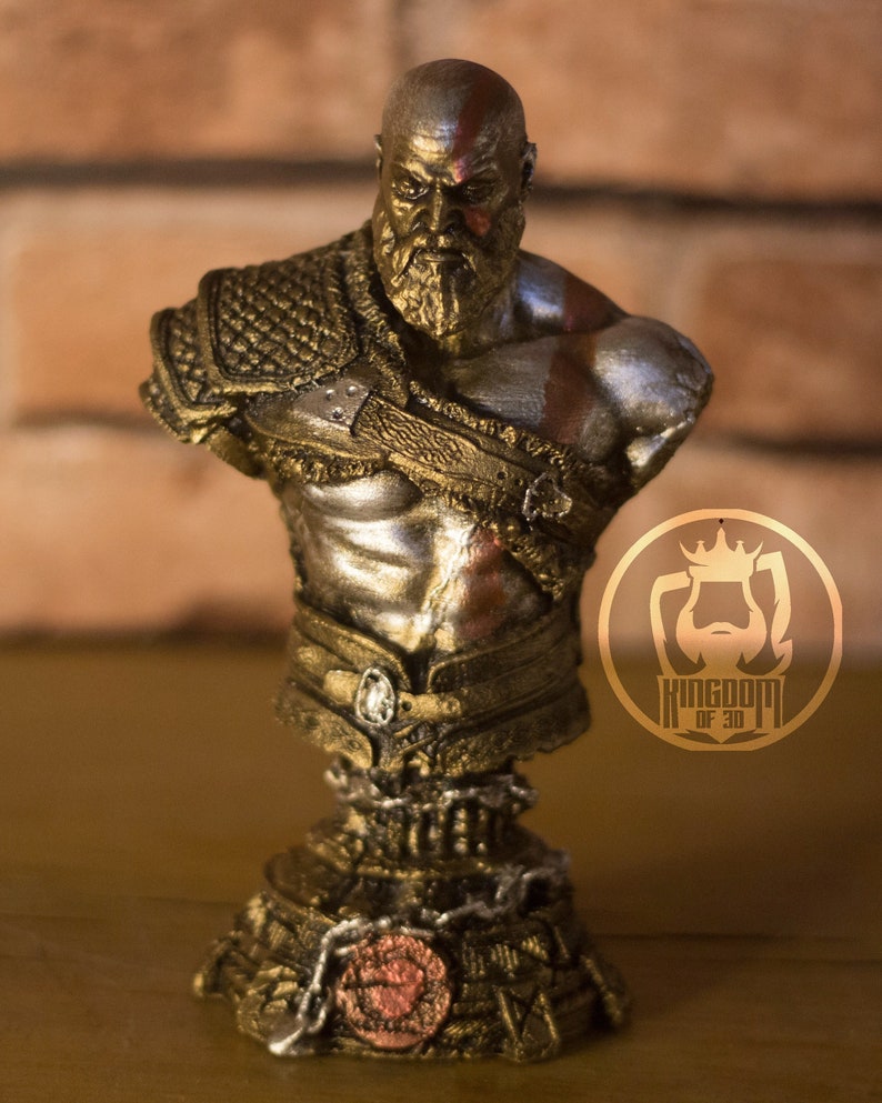 May include: A detailed bust of a warrior figure, featuring a bald head with a red stripe, a full beard, and intricate armor. The sculpture is predominantly gold and silver, with a detailed base and the text "KINGDOM OF 3D" on a circular emblem.