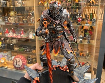 May include: A detailed figurine of a knight in dark armor with fiery orange accents. The knight holds a large, red, flaming sword and stands on a decorative base with autumn leaves. The figurine is a collectible item.