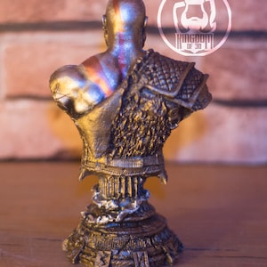 May include: A metallic gold bust of a bald warrior with a red stripe on his head and shoulder armor. The bust is on a decorative pedestal. The logo "KINGDOM OF 3D" is visible in the background.