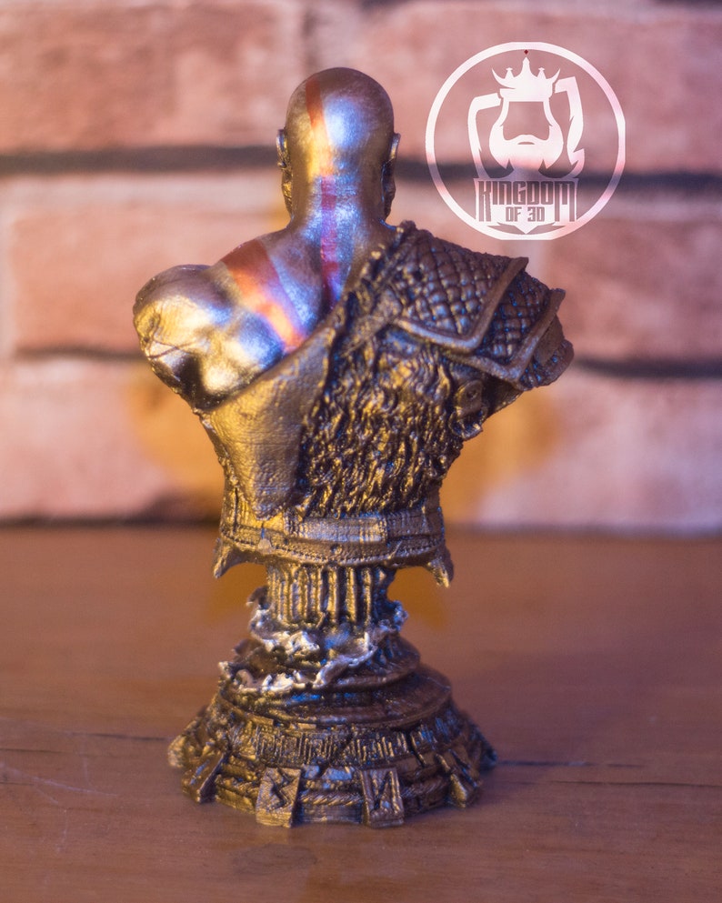 May include: A metallic gold bust of a bald warrior with a red stripe on his head and shoulder armor. The bust is on a decorative pedestal. The logo "KINGDOM OF 3D" is visible in the background.
