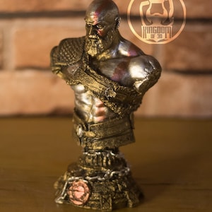 May include: A detailed bust of a bearded warrior with a stern expression, featuring gold and silver accents. The figure has a red painted mark on the head and chest. The base is intricately designed with chain details and a circular emblem.
