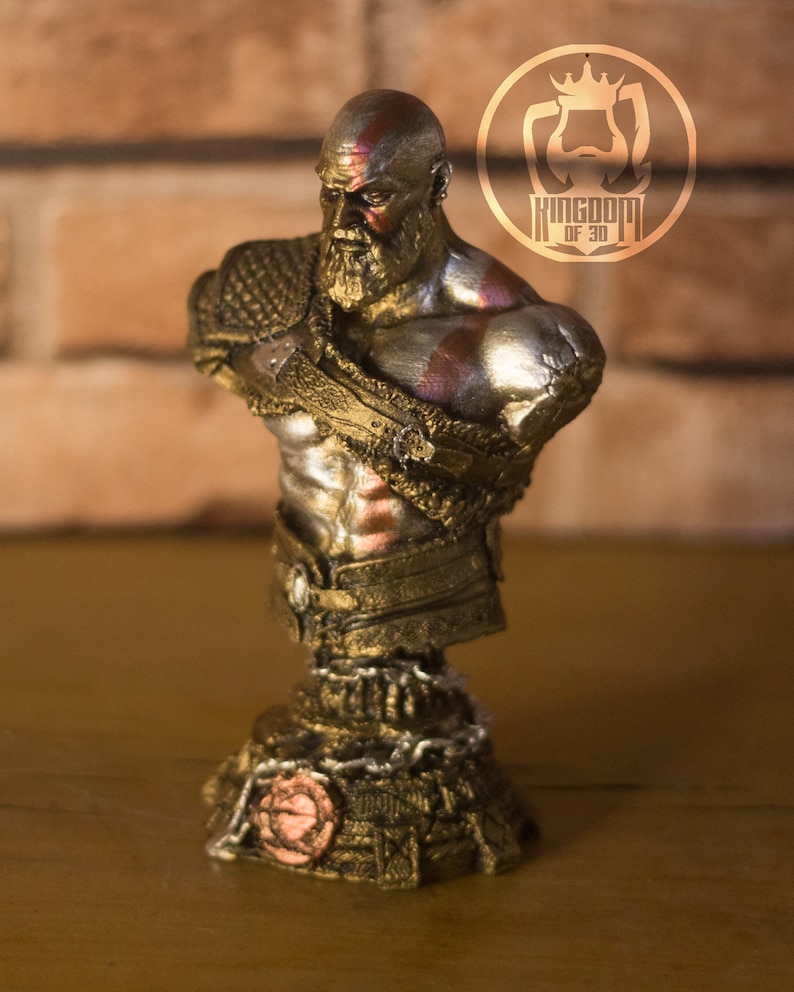 May include: A detailed bust of a bearded warrior with a stern expression, featuring gold and silver accents. The figure has a red painted mark on the head and chest. The base is intricately designed with chain details and a circular emblem.