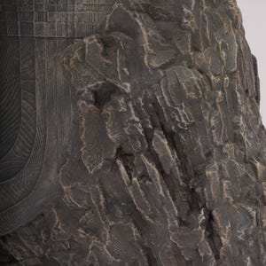 May include: A dark brown sculpture featuring a textured, rugged rock-like formation on the right side, contrasted with a smooth, curved section on the left that displays a detailed, grid-like pattern of small, rectangular, etched indentations.