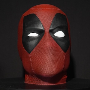 Puede incluir: Una máscara de Deadpool roja y negra. La máscara presenta una superficie texturizada, similar a la tela. Las aberturas de los ojos son blancas y las áreas circundantes son negras. La máscara está sobre un fondo oscuro.