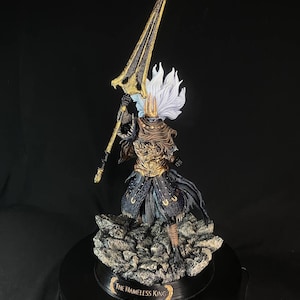 May include: A detailed figurine of The Nameless King from Dark Souls III. The figure is posed holding a spear with a golden blade and a black shaft. The figure is wearing a black and gold armor with a white cape. The figure is standing on a rocky base with a black circular base.