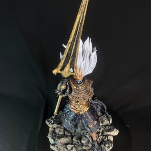 May include: A detailed figurine of the Nameless King from Dark Souls III. The figure is posed in a dynamic stance, holding a spear with a golden blade. The figure is wearing a black and gold armor with a white cape. The figure is standing on a rocky base with the words "The Nameless King" inscribed on the base.