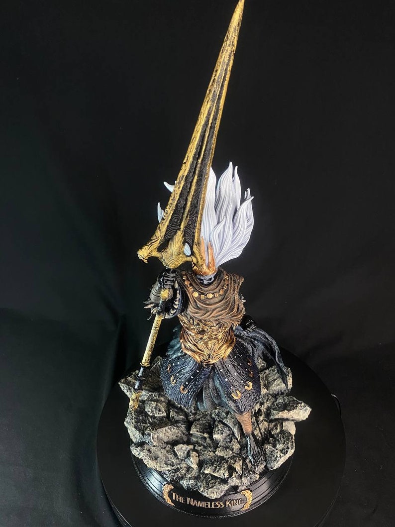 May include: A detailed figurine of the Nameless King from Dark Souls III. The figure is posed in a dynamic stance, holding a spear with a golden blade. The figure is wearing a black and gold armor with a white cape. The figure is standing on a rocky base with the words "The Nameless King" inscribed on the base.