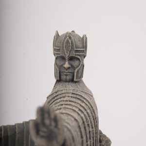 May include: A gray, textured figurine of a figure wearing a detailed helmet and a long, layered robe. The figure is depicted with one arm extended forward, palm facing outward, showcasing a weathered, stone-like finish and intricate carvings.