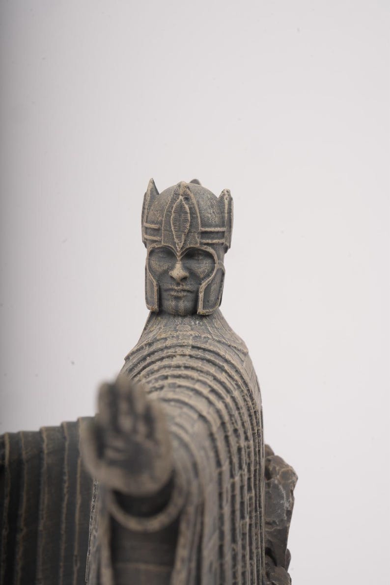 May include: A gray, textured figurine of a figure wearing a detailed helmet and a long, layered robe. The figure is depicted with one arm extended forward, palm facing outward, showcasing a weathered, stone-like finish and intricate carvings.