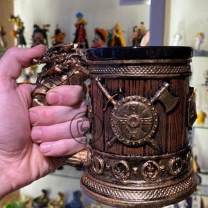 May include: A bronze-coloured Viking-themed mug. The mug has a wooden barrel design with a shield, crossed swords, and an axe. The handle is sculpted with a dragon motif. The top of the mug is black.