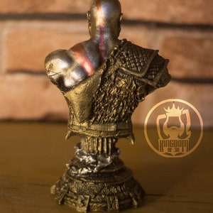 May include: A bronze-colored bust of a bald figure, viewed from the back, with red and silver markings on the head and back. The figure wears ornate armor with a textured design. The base of the bust has a detailed, layered appearance. The logo "KINGDOM OF 3D" is visible.