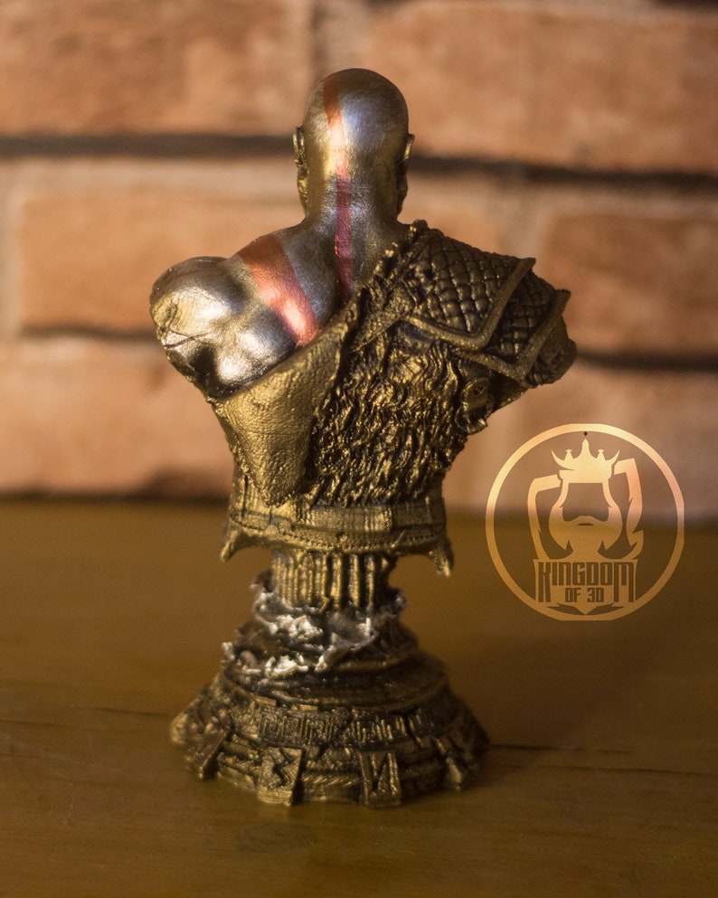 May include: A bronze-colored bust of a bald figure, viewed from the back, with red and silver markings on the head and back. The figure wears ornate armor with a textured design. The base of the bust has a detailed, layered appearance. The logo "KINGDOM OF 3D" is visible.